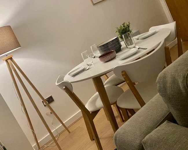 A modern dining room features a round table set for a meal, surrounded by white chairs, with a floor lamp and a couch nearby.