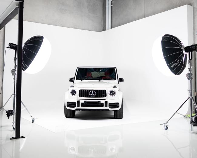 A white Mercedes-Benz G-Class is positioned in a well-lit studio with large softbox lights on either side and a seamless white backdrop.
