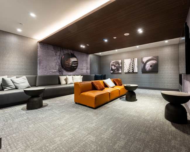 The image shows a modern lounge area featuring a gray sofa, an orange seating unit, two small black side tables, and framed artwork on the walls.