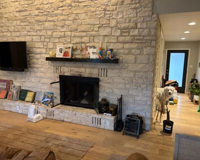 The image shows a room with a stone wall featuring a fireplace, a television mounted above, and an adjacent area with plants and various items.