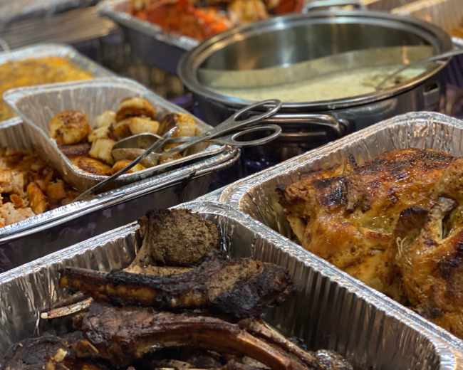 A variety of cooked dishes served in aluminum trays, including roasted meats, seafood, and sides.