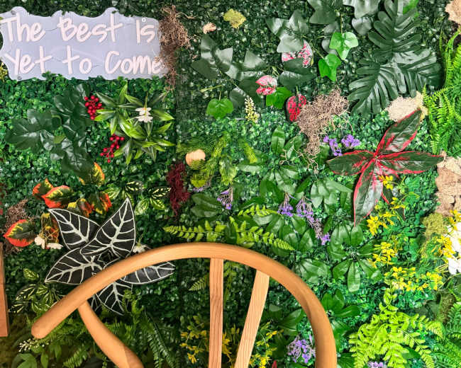 The image shows a vibrant wall covered in various artificial plants and leaves, along with a wooden chair in the foreground.