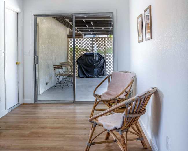 A brightly lit room features two wooden chairs facing a sliding glass door that opens to a covered patio area.