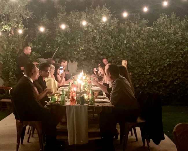 A long outdoor dining table is set with candles and surrounded by people enjoying a meal under string lights.