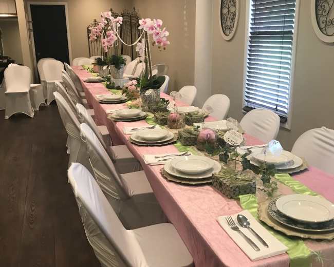 A long dining table is set with pink and green tablecloths, white chairs, and floral decorations, arranged for an elegant gathering.