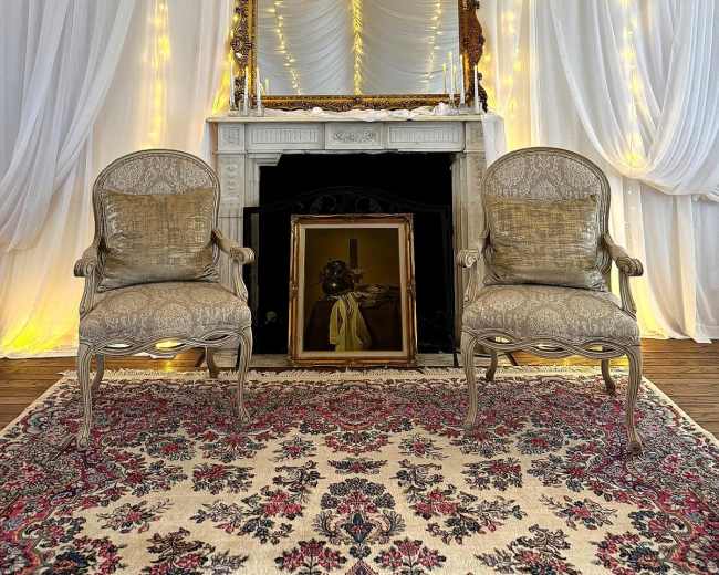 The scene features an elegant room with two ornate chairs facing a vintage mirror above a decorative fireplace, all set against draped white fabric and soft, warm lighting.