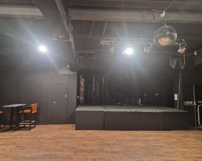 The image shows a dimly lit performance stage with a disco ball and a seating area featuring high tables and chairs.