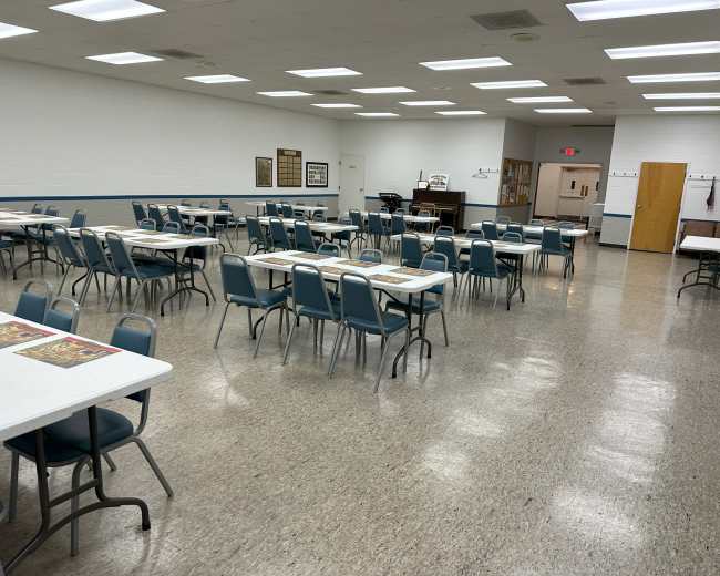 A large, empty community hall features rows of tables and chairs arranged in a grid pattern under bright overhead lights.