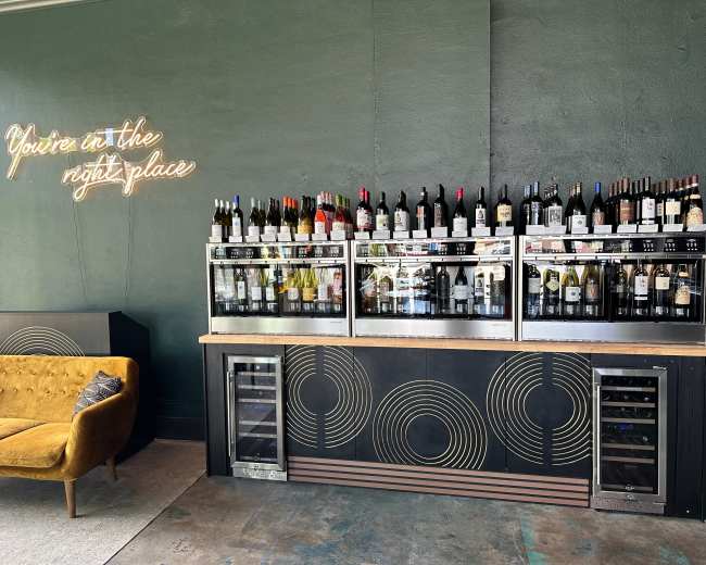 A wine display with various bottles is set against a dark green wall, accompanied by a neon sign that reads "You're in the right place."