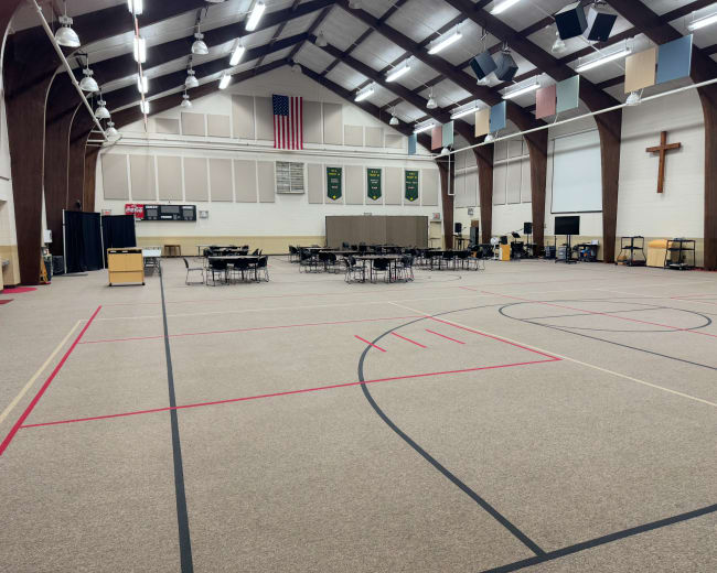 The image shows a large indoor gymnasium with a wooden-beamed ceiling, rows of chairs set up at tables, and a cross on the wall.