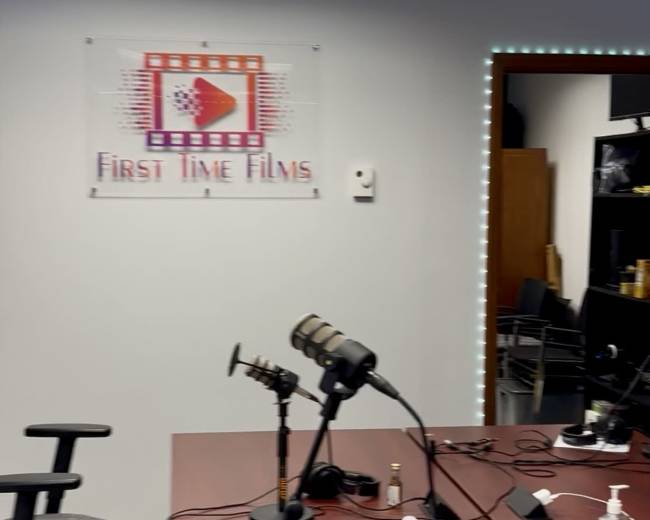 The image shows a film studio or podcast recording space with a table, microphones, and a sign for "First Time Films" on the wall.