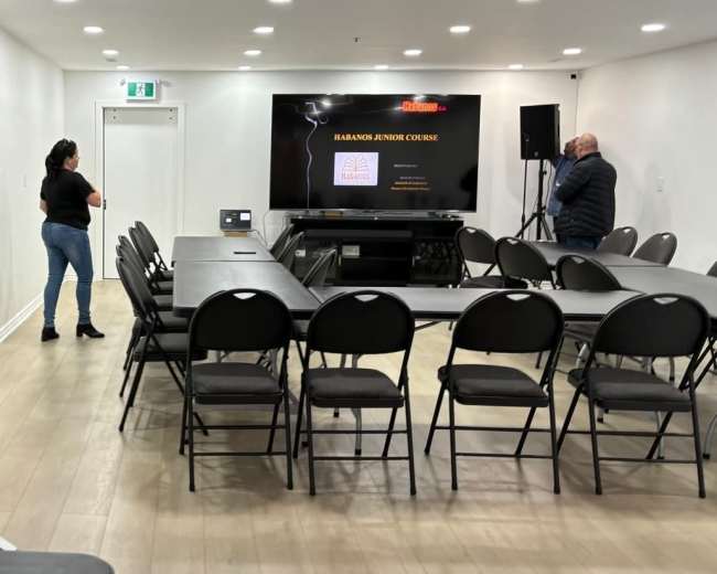 A small, modern conference room is set up with rows of chairs facing a screen displaying "HUMANO ATINO COURSE" while two people stand in the background.