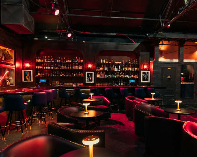 A dimly lit bar features a long shelf of bottles in the background, with small tables and dark chairs arranged in a cozy layout.