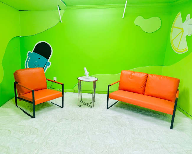 The image shows a small, brightly colored room with green walls, two orange sofas, and a round white table.