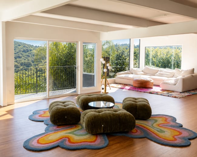The image shows a spacious living room with large windows overlooking a green hillside, featuring a colorful area rug, modern furniture, and natural light.