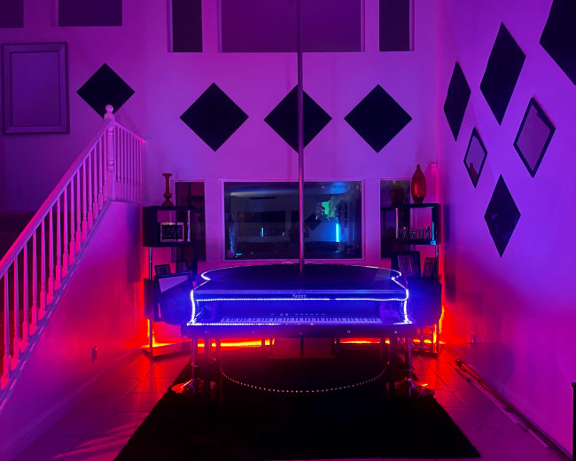 A grand piano with neon blue and red lights in a room featuring a pole and diamond-shaped wall decorations.