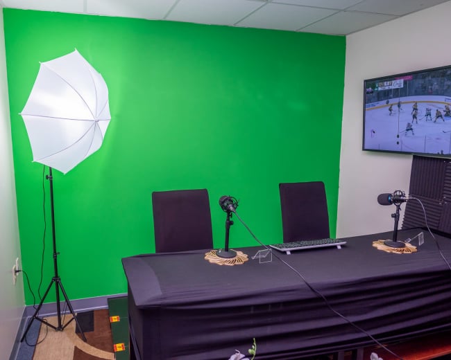 A small studio with a green screen wall, two chairs at a table, microphones on stands, and a monitor displaying a sports game.
