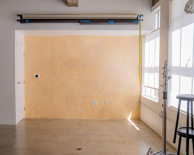 The image shows a sparsely furnished room with a smooth, textured orange wall, concrete flooring, and large windows that allow natural light to enter.