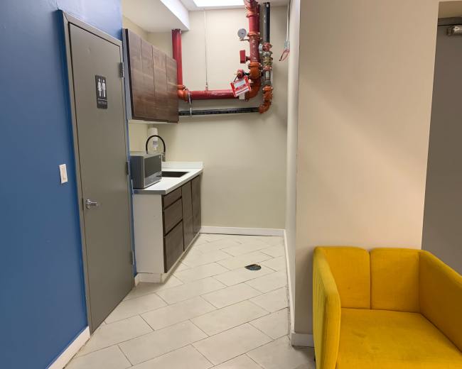 The image shows a small kitchenette area with a sink and cabinets, adjacent to a hallway featuring a yellow couch and exposed plumbing.