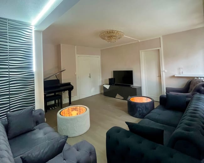 The image shows a modern living room featuring a piano, a television, and two circular coffee tables with glowing inserts, surrounded by plush seating.