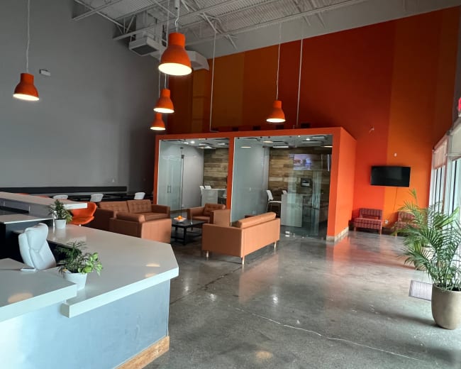 The image shows a modern reception area with a front desk, orange pendant lights, and a seating area featuring brown sofas and glass-walled conference rooms.