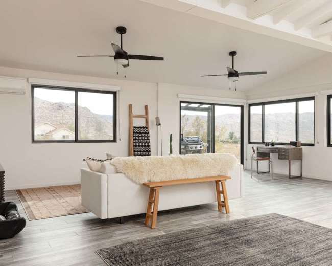 The image shows a modern, open-concept living space with large windows overlooking a mountainous landscape, featuring a white sofa, a wooden bench, and a desk in the corner.