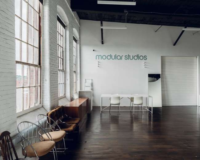 The image shows a spacious, brightly lit studio with large windows, a white wall featuring the text "modular studios," and a minimalist furniture arrangement including several chairs and a table.