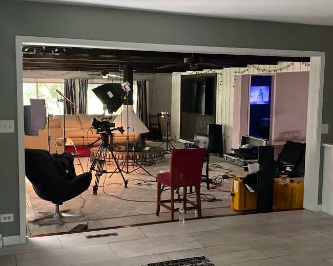 A film set is arranged in a living room, featuring cameras, lighting equipment, and chairs, with scattered production gear on the floor.
