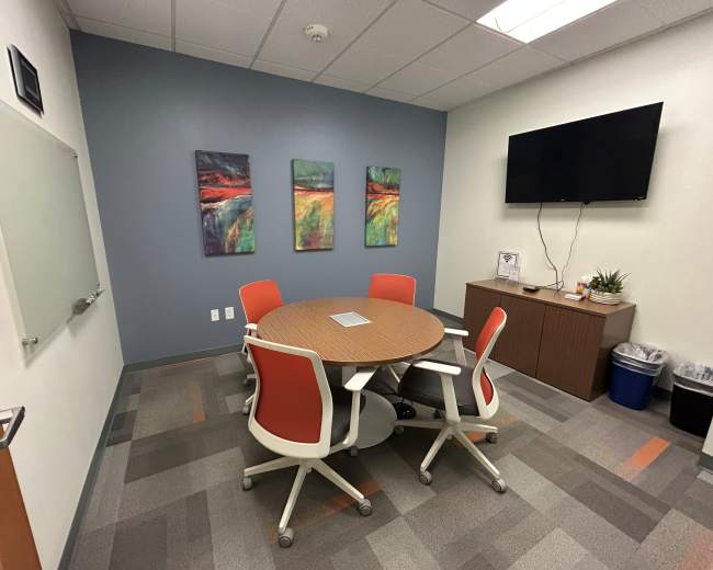 The image shows a small conference room featuring a round table with five chairs, three abstract paintings on the wall, a television mounted in one corner, and a cabinet with a plant and waste bin.