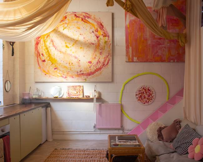 The image shows a cozy, brightly lit room featuring colorful abstract paintings on the walls, a simple kitchen area, and a small seating area with a couch.