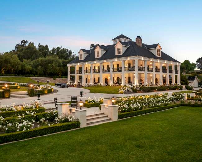 A large, elegant mansion with a white exterior and multiple balconies is surrounded by manicured gardens and landscaped pathways.