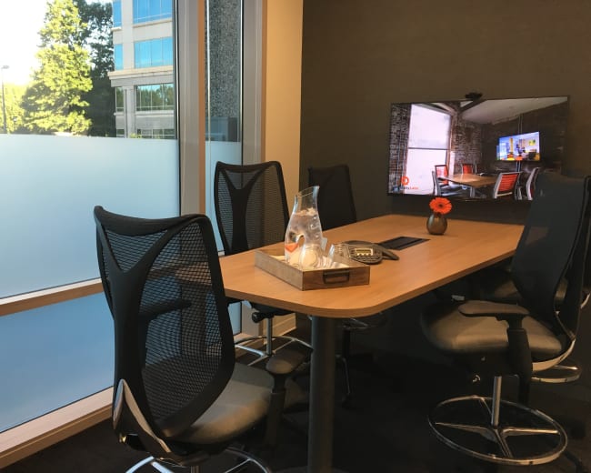 A modern conference room features a wooden table surrounded by ergonomic chairs and a wall-mounted television displaying a presentation.
