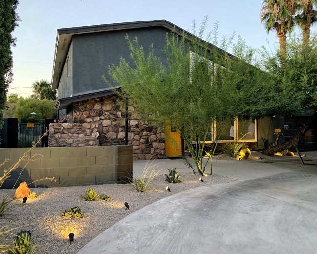 The image shows a modern house with a stone facade, surrounded by low-maintenance landscaping, and illuminated by landscape lighting in the evening.