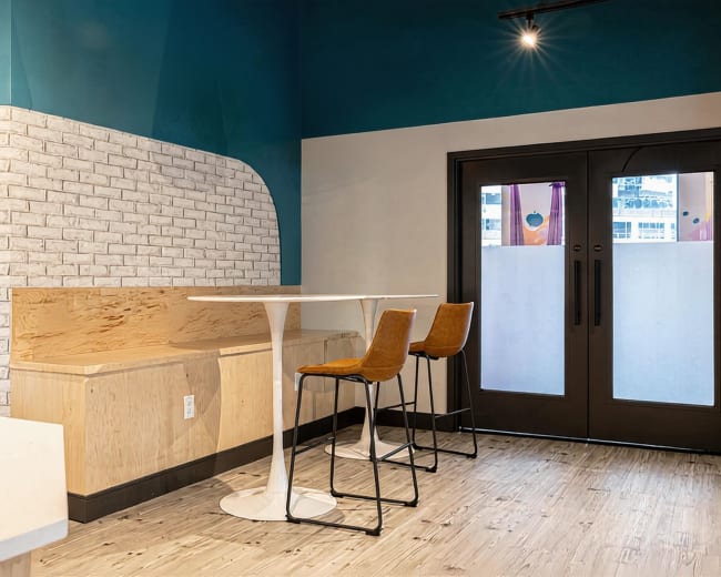 The image shows a modern interior space featuring a light wooden bench, a circular white table, and three brown bar stools, with a blue and white color scheme and large double doors.