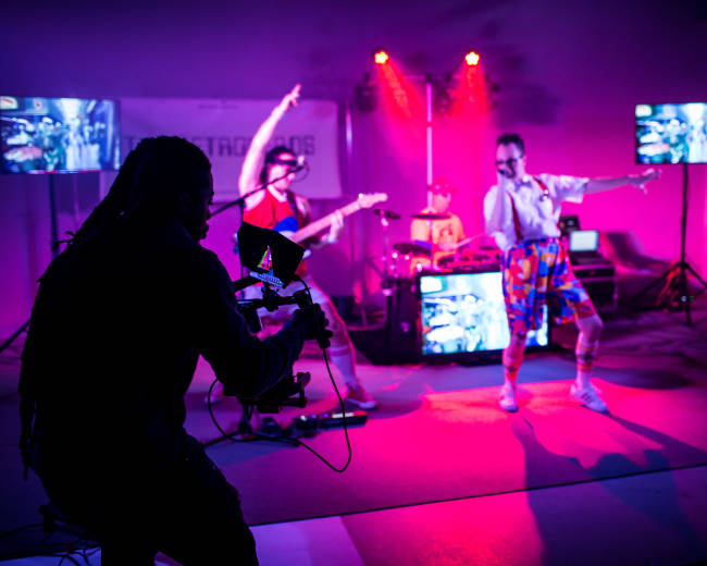 A camera operator captures a band performing on stage, surrounded by colorful lights and video screens displaying their performance.