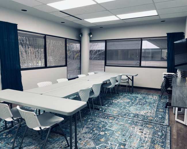 A conference room features multiple tables arranged in a U-shape, surrounded by chairs, with large windows allowing natural light to enter.