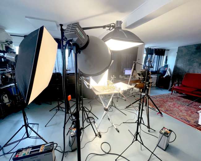 The image shows a photography studio setup with multiple light sources, a table arranged with equipment, and a red sofa in the background.