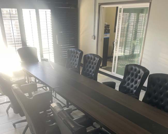 A long wooden conference table with black leather chairs is positioned in a brightly lit room with large windows.