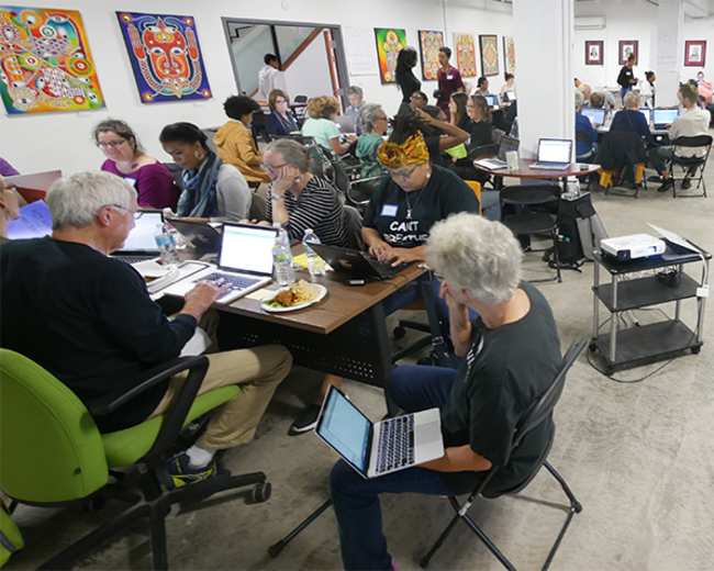 A large group of people working individually and in small clusters at tables in a bright room filled with artwork.