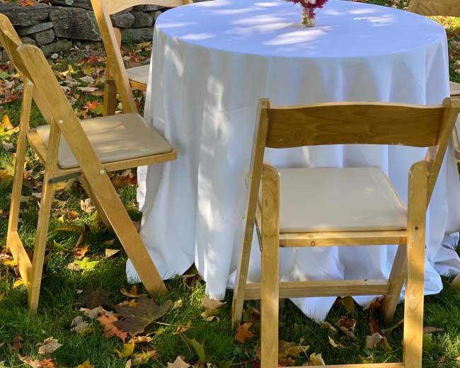 A round table covered with a white tablecloth is set with a small floral centerpiece, surrounded by three wooden chairs on a lawn with autumn leaves.