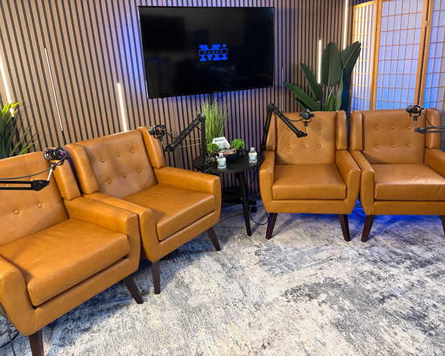 The image shows a cozy sitting area with three brown leather chairs, a small round table, and a backdrop featuring a TV screen and plants.