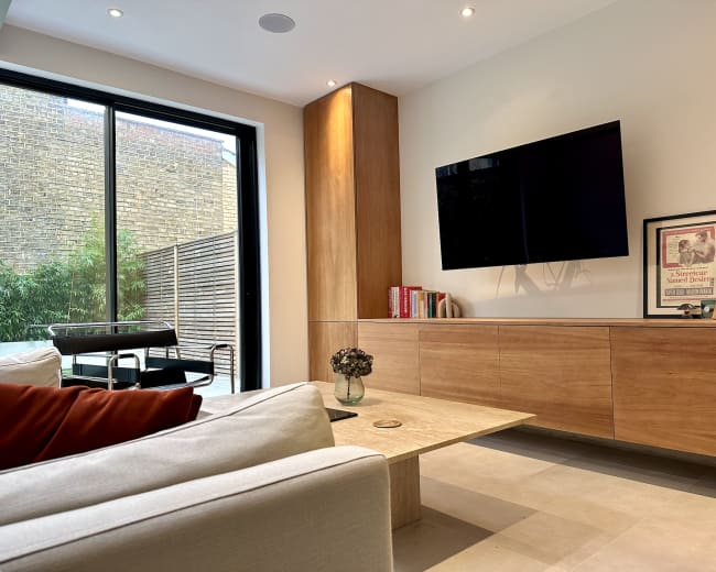 The image depicts a modern living room with a television mounted on the wall, a wooden coffee table, a sofa with cushions, and a view of a small outdoor space visible through large glass doors.