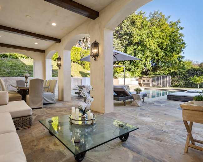 A patio area featuring a glass coffee table, comfortable seating, and a view of a swimming pool and landscaped greenery outside.