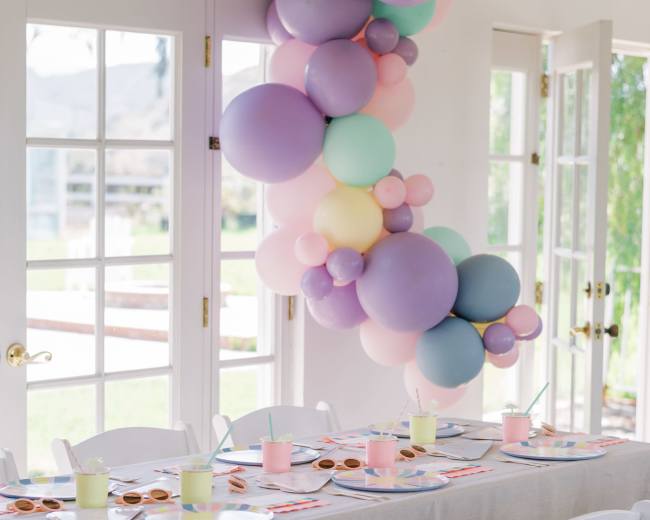 A table is set with pastel-colored plates and cups, decorated with a balloon garland, in a bright room with large windows.