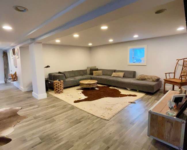 A spacious basement living area features a gray sectional sofa, a round coffee table, and a decorative area rug on a wooden floor, illuminated by recessed lighting.