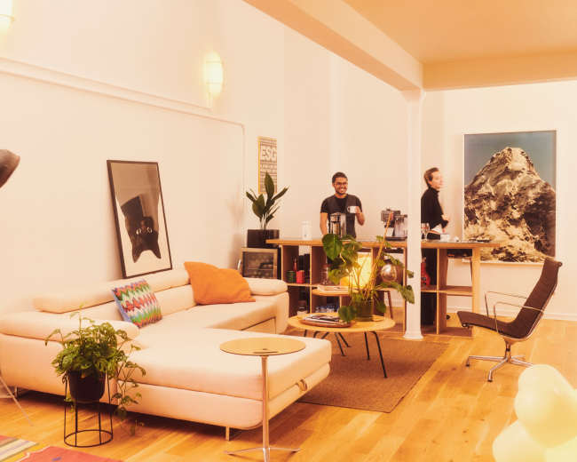 The image shows a modern office space featuring a light-colored couch, a wooden desk with two people, and a large mountain artwork on the wall.