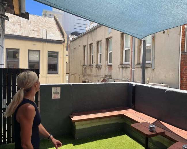A woman stands on a small rooftop terrace with artificial grass and wooden benches, overlooking a narrow alley between buildings.