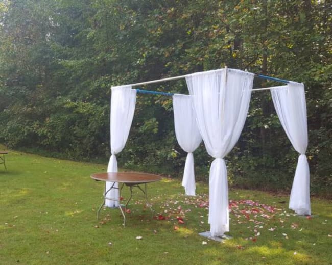 A wedding gazebo draped in white fabric stands on a grassy area surrounded by scattered rose petals and several round tables.
