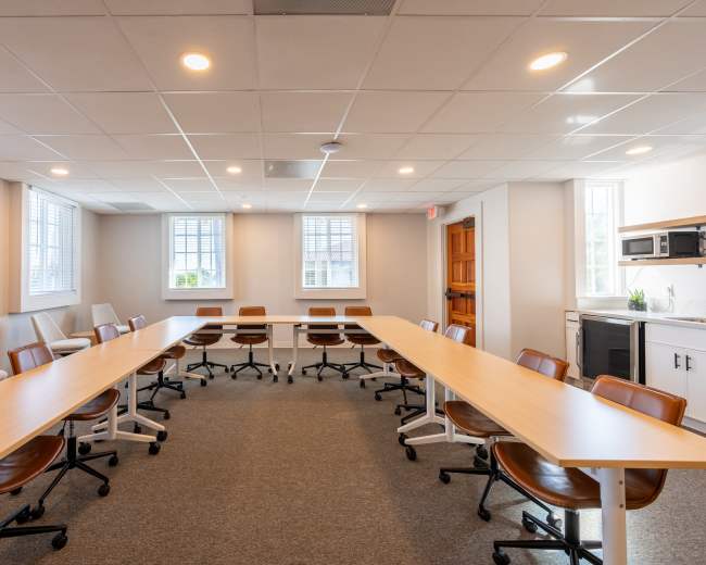 A modern conference room features a long table surrounded by ergonomic chairs, with windows providing natural light and a kitchenette in the corner.