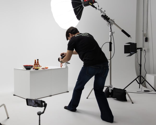A photographer wearing a black shirt captures an image of food on a table in a studio setting with multiple lighting sources.